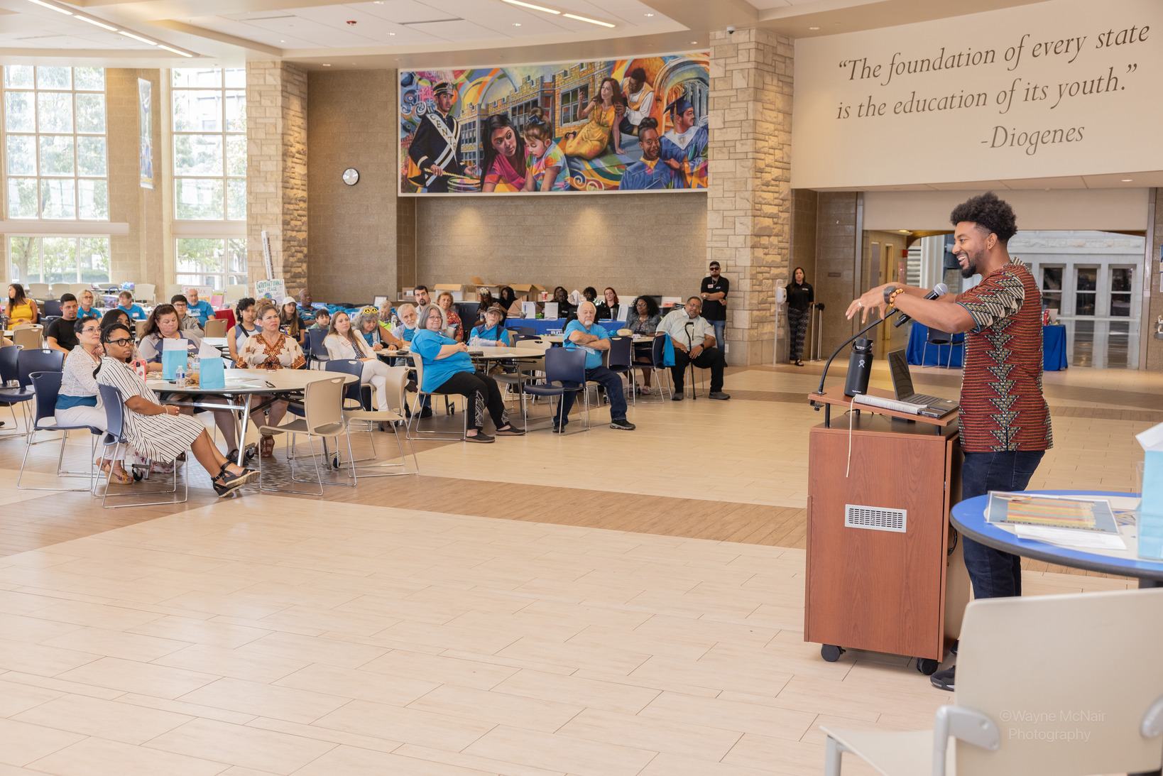 Ernest Crim III speaking from a podium to a seated audience in a school atrium